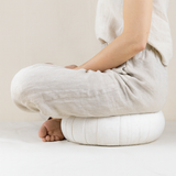 Femme assise en position de lotus sur un zafu rond en lin écru, vêtue d’une tenue en lin naturel, dans un décor épuré et apaisant – Atelier Cocon