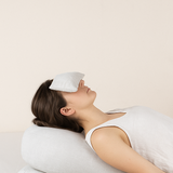 Femme allongée dans une posture de relaxation restorative yoga, le dos soutenu par un bolster en lin greige, un coussinet pour les yeux en lin posé sur son visage, ambiance zen et lumineuse – Atelier Cocon