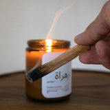 Bougie naturelle en cire de soja allumée sur une table en noyer, avec un bâton de palo santo fumant tenu à la main.